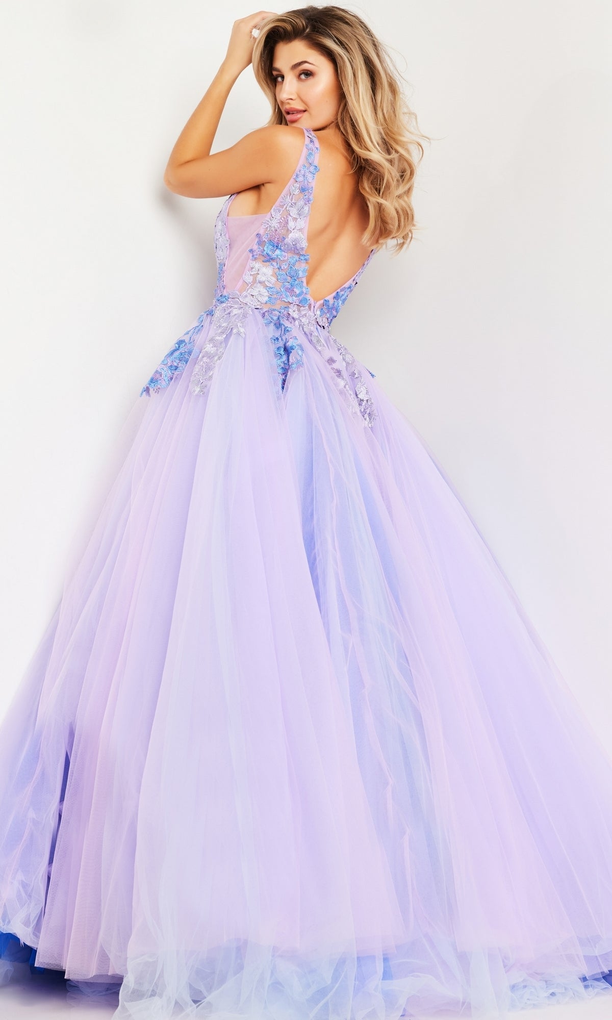 A woman poses against a white background, smiling over her shoulder and showcasing the back of the Jovani Lilac Purple Long Prom Ball Gown 24602 featuring floral appliqué details and an elegant A-line silhouette.